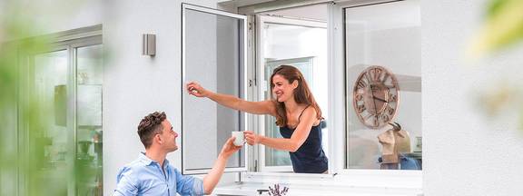 Frau reicht Mann auf Terrasse eine Tasse Kaffee durch Insektenschutzfenster