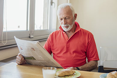 Älterer Herr sitzt zum Frühstück am Tisch neben einem Fenster und liest Zeitung