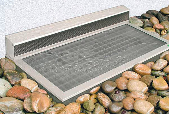 Eine vom Regen benetzte RESI-Lichtschachtabdeckung ist in einem stilvollen Steinbeet auf einem Kellerschacht montiert.