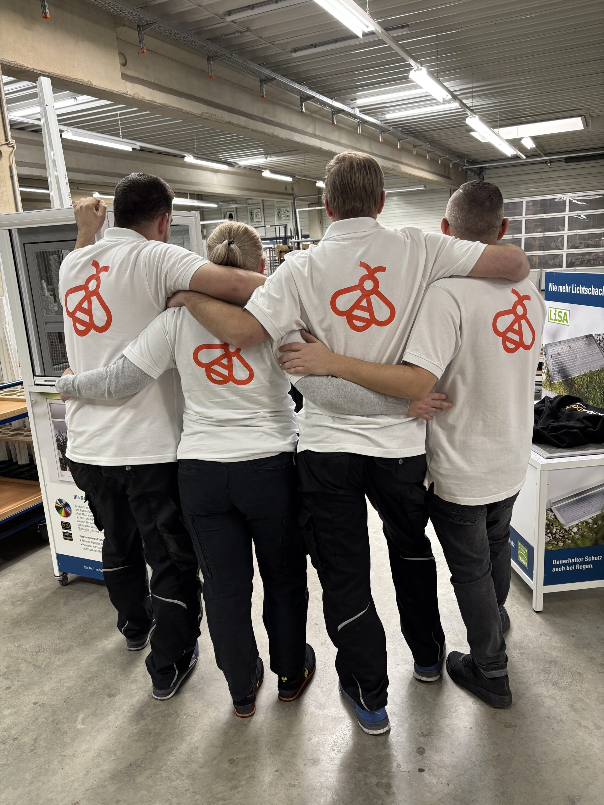 Vier Mitarbeitende von Schulz-Dobrick stehen Arm in Arm nebeneinander. Sie stehen mit dem Rücken zur Kamera. Alle tragen ein weißes Shirt, auf dem hinten ein großes oranges Bienen-Icon zu sehen ist.