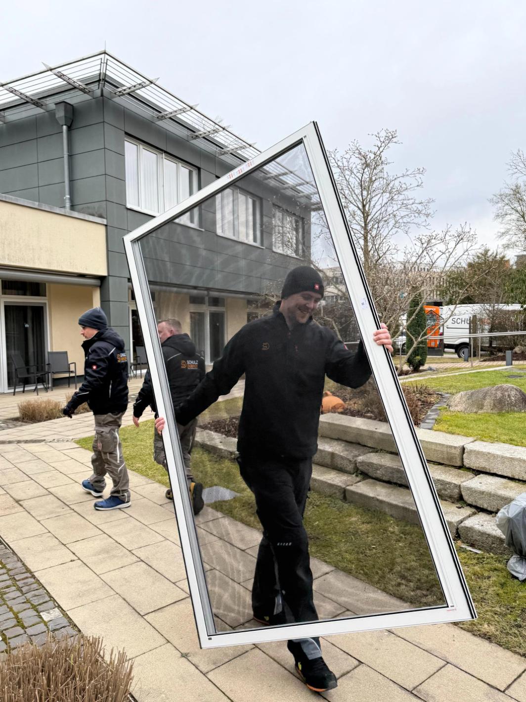 Drei Monteure gehen aneinander vorbei. Zwei gehen in Richtung der Montage-Fahrzeuge. Der dritte trägt einen Insektenschutzgitter mit weißem Rahmen.