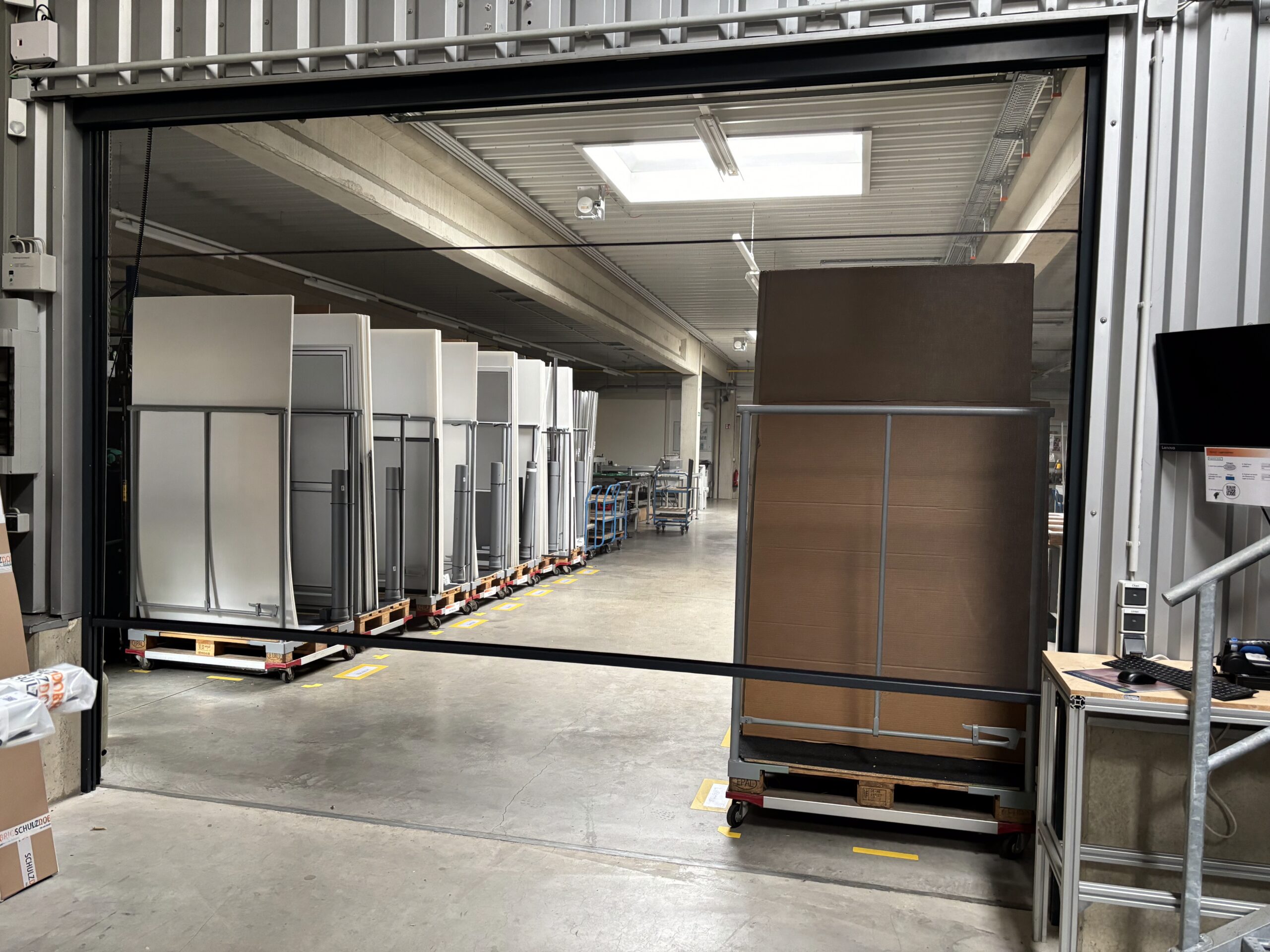 Elektrorollo in einem breiten Durchgang einer Produktionshalle. Schwarze Rahmen und durchsichtiges Insektenschutzgewebe.